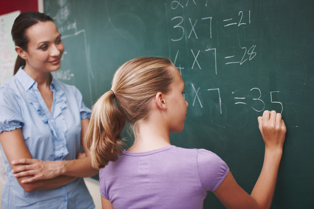 A student writing on the blackboard in a maths class, watched by a smiling teacher
