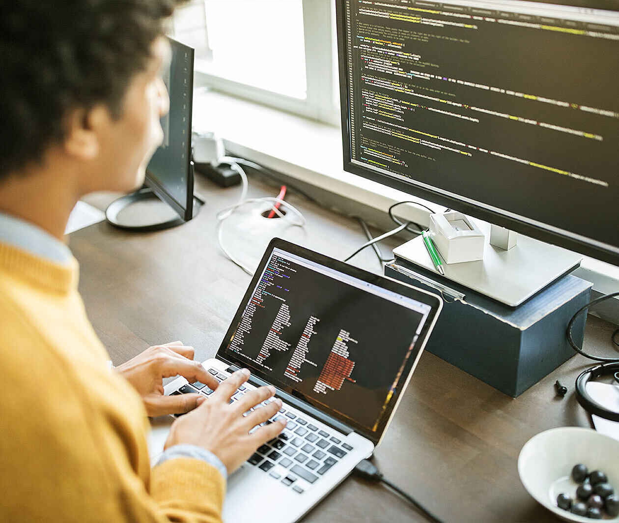 Man wearing a yellow jumper looking at two screens of code.