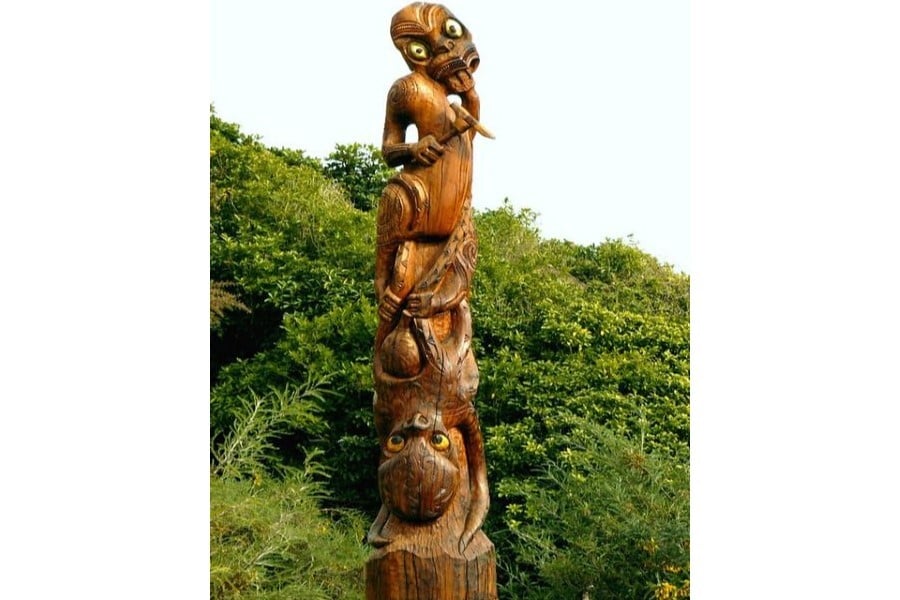 A photograph of a carved wooden pillar surrounded by lush green bush. The traditional Māori carvings depict a stylized Māori figure at the top, his tongue out and a weapon in hand, atop an octopus, whose tentacles reach up to grab him. Both figures are co