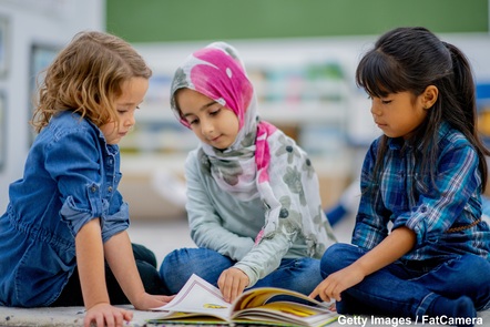 A group of three young female pupils. The pupil on the left is resting on her hands, the pupil in the middle is turning a page in a book they are all looking at, and the pupil on the right is pointing to the open page.