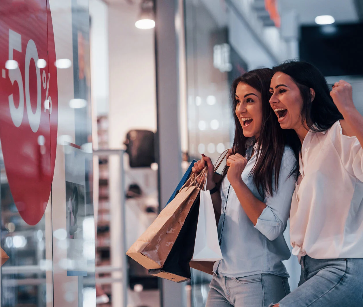 Image of 2 cheerful females shopping in a mall and excited about a 50% off sale sign in a window