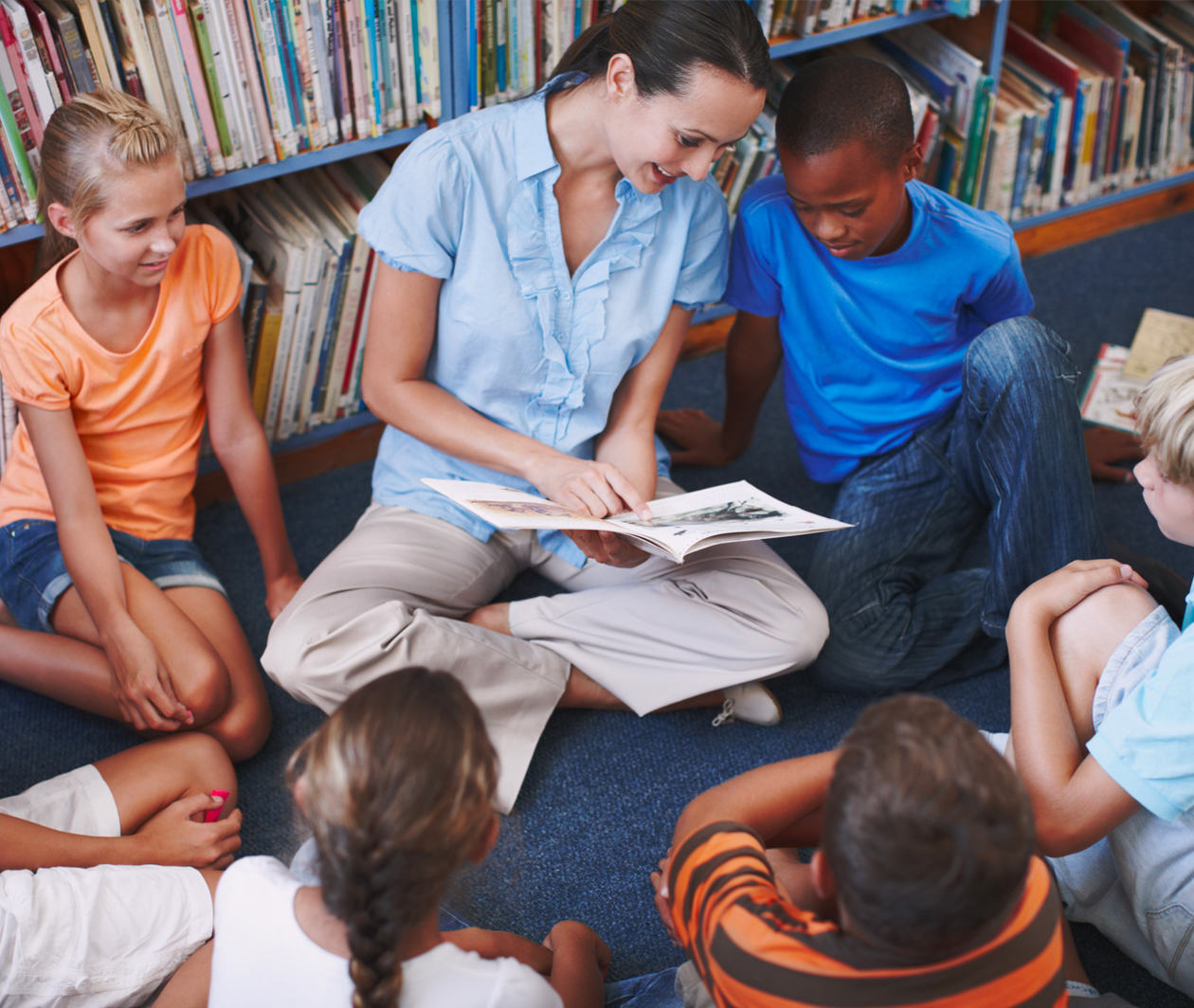 Woman reads picturebook to a group of children sitting in a circle in a library setting. The woman points at the pictures and text while the children look on with apparent interest.