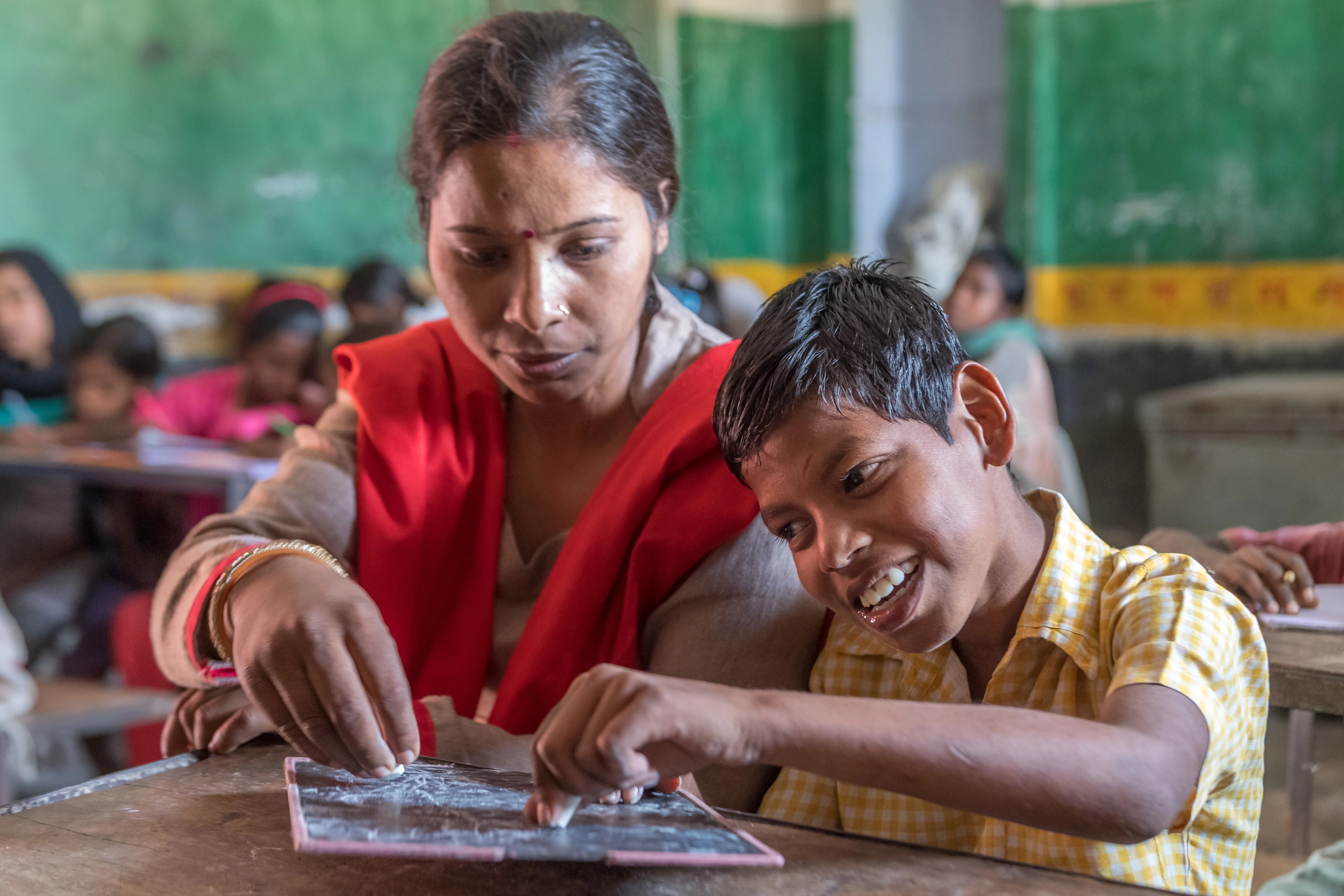 A boy with cerebral palsy writing on a chalk board alongside his teacher in school