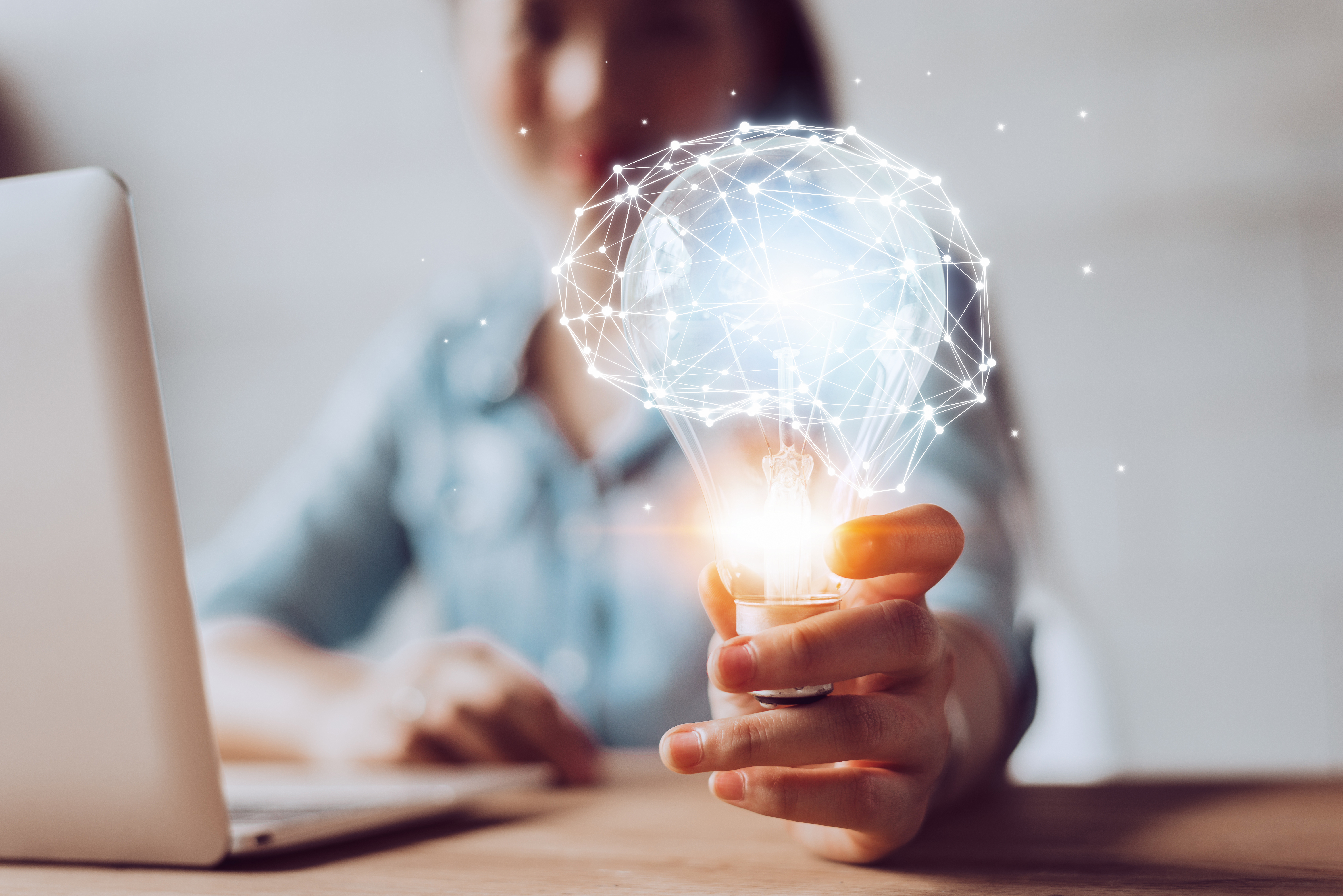 Woman sitting at her laptop holds a lightbulb with lights making the shape of a brain around it.