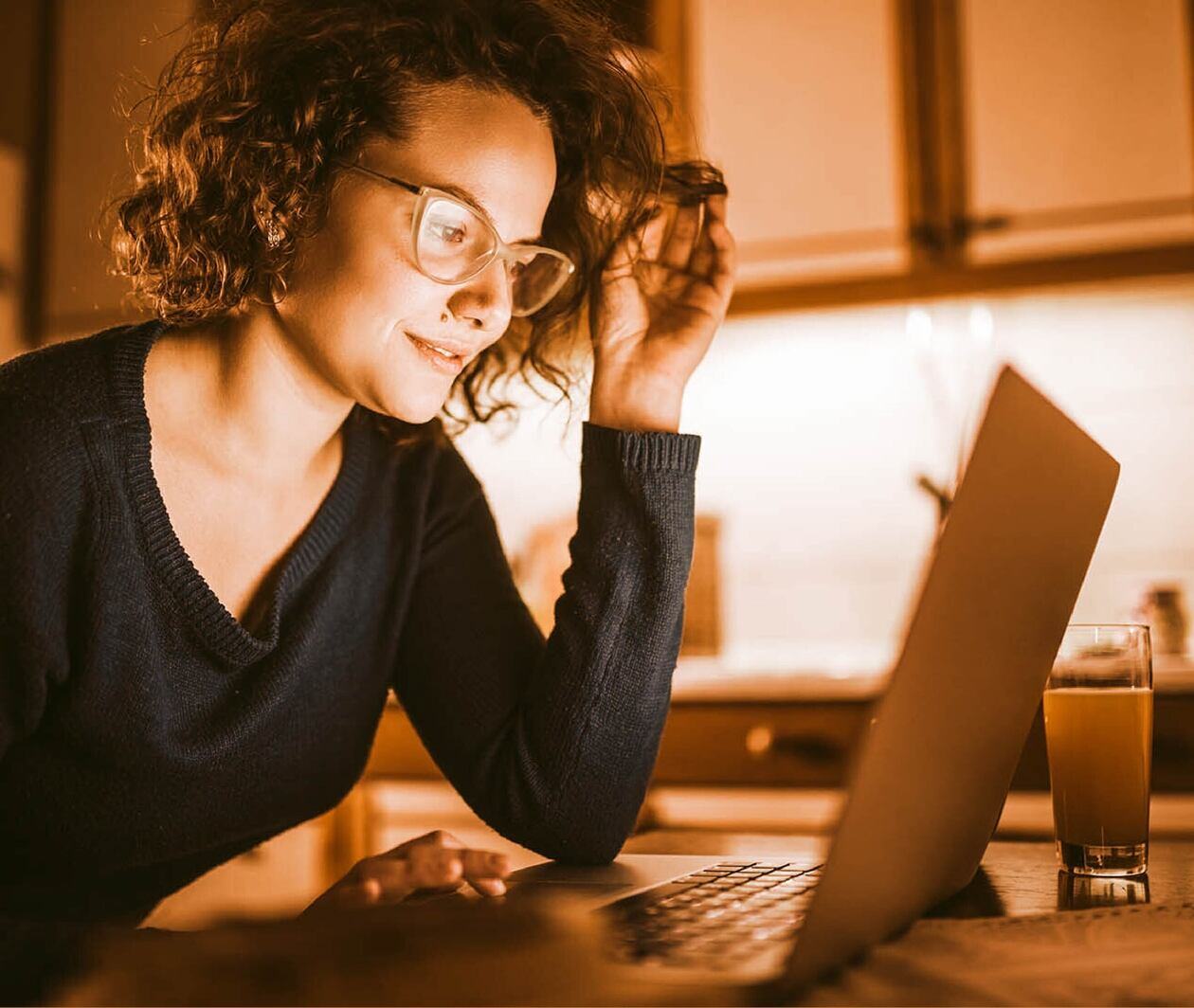 A woman in a black jumper and glasses is on a laptop in her kitchen. 