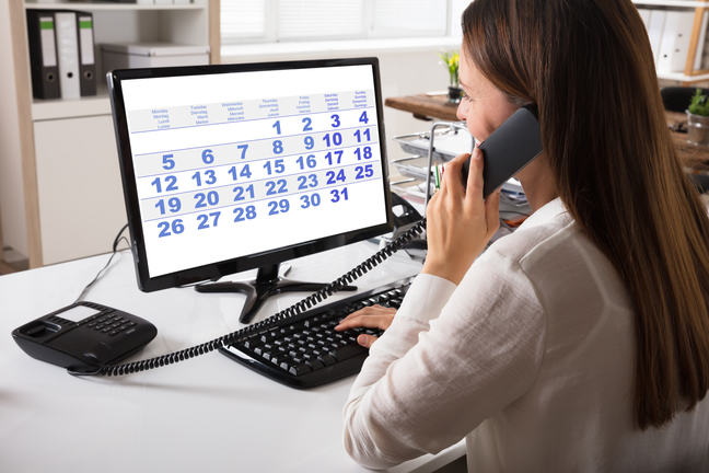 A woman on a telephone looking at a computer screen with a calendar on it.