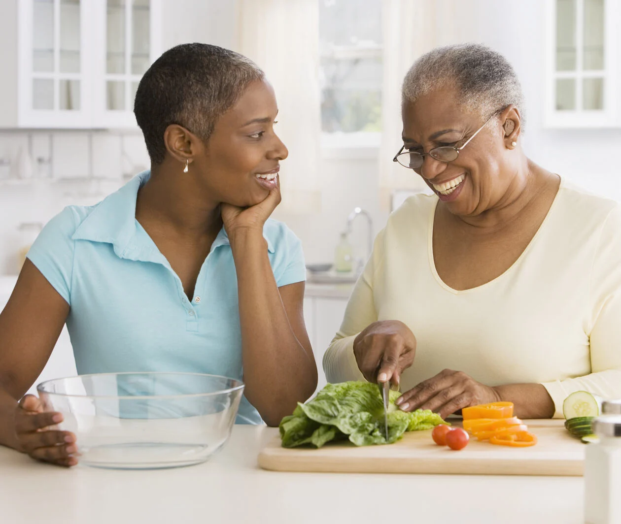 A young woman on the left holds a glass bowl while speaking to an older woman on her right chopping vegetables on a chopping board.