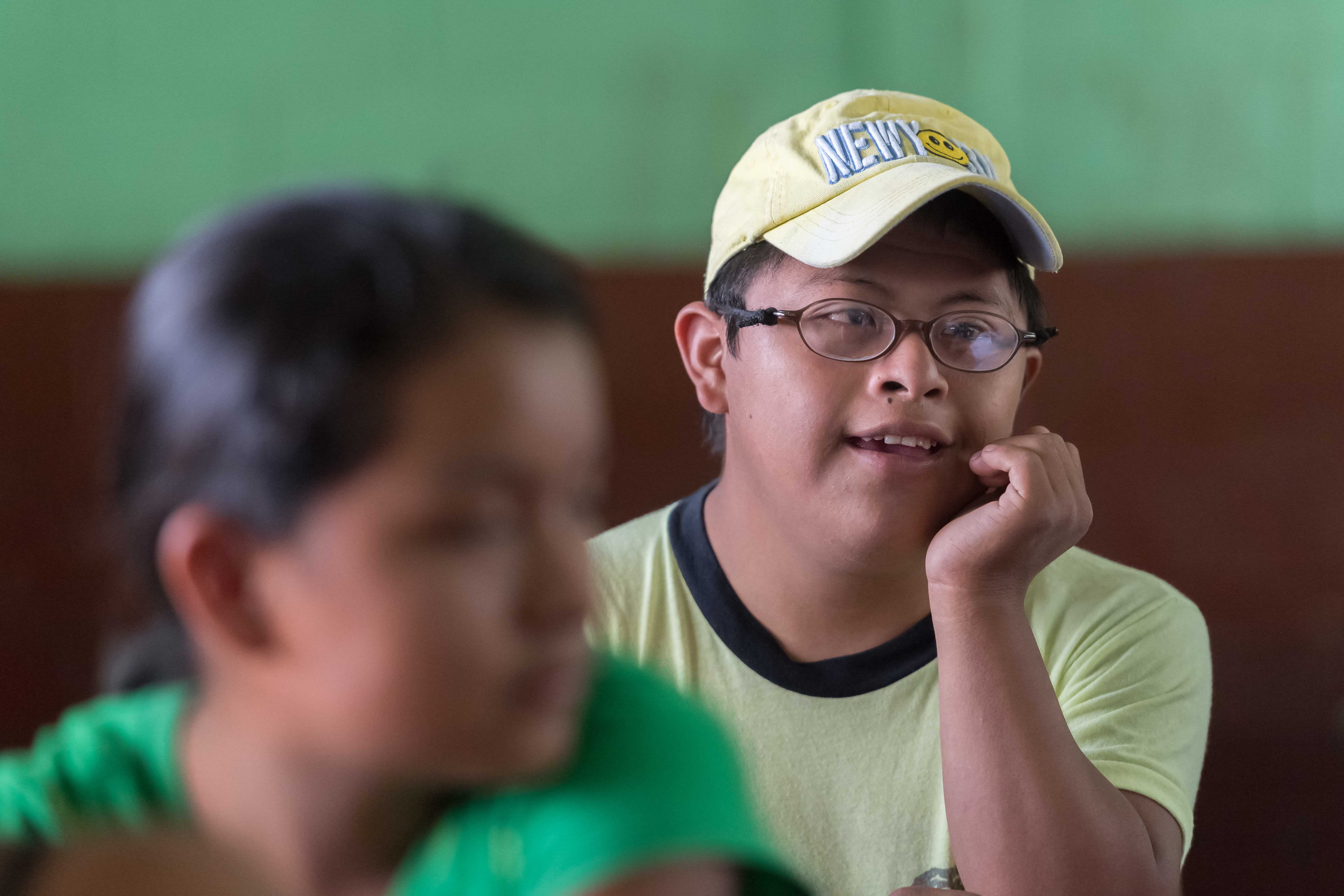 A teenage boy with Down syndrome sitting in a school classroom. He is wearing a baseball hat and glasses and leaning on one hand.