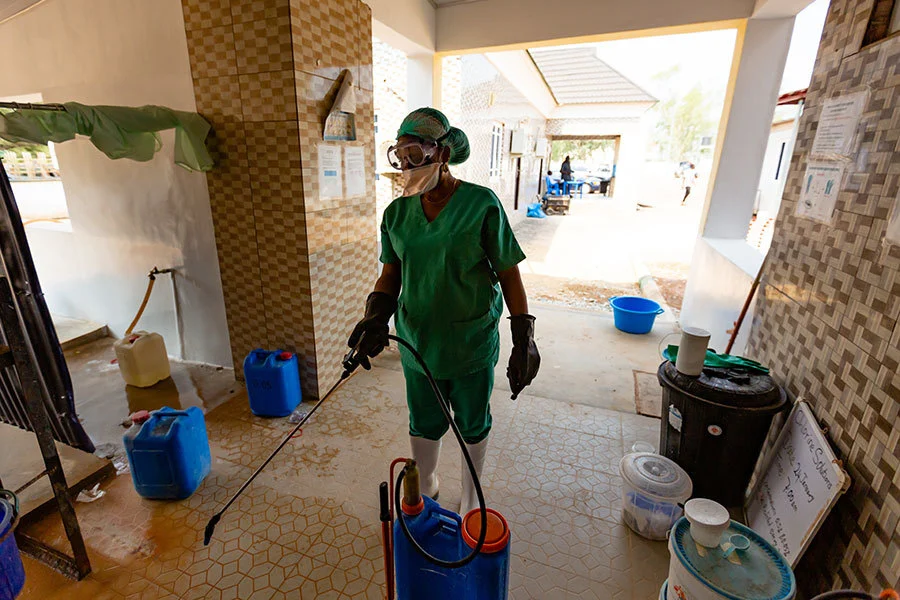 A nurse wearing a scrub suit, gloves, mask, goggles and hairnet is holding decontamination equipment which has a long tube with a nozzle and a blue container. She is spraying the floor of the building. There are containers at her side.