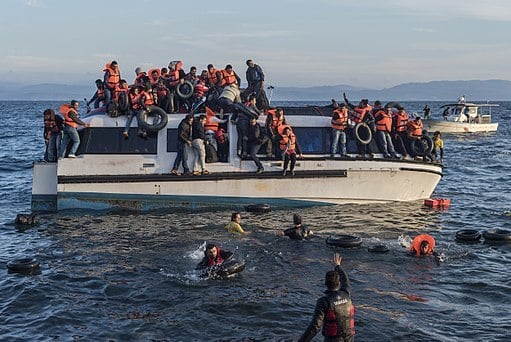 An overcrowded boat transporting lots of refugees. A number of them are in the water.