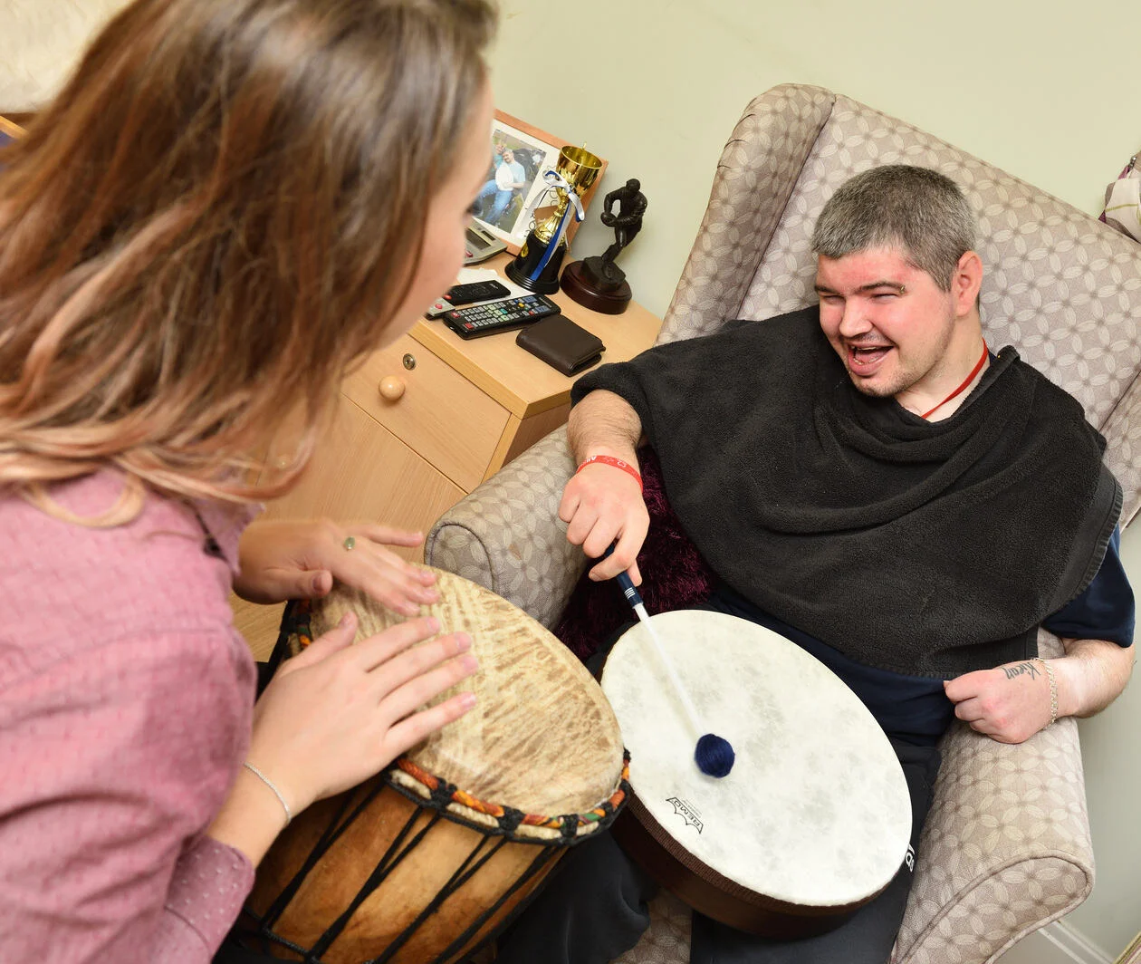 A female music therapist has her back to the camera playing a hand drum, she is facing a young man playing a smaller drum with a beater