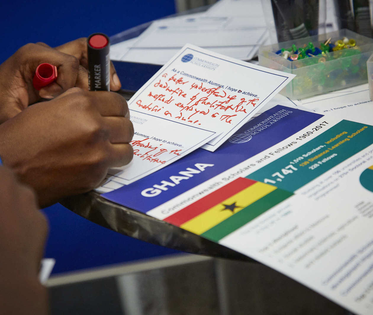 Close-up image of a Scholar writing on a comment card at a CSC event with information sheet about Ghana nearby.