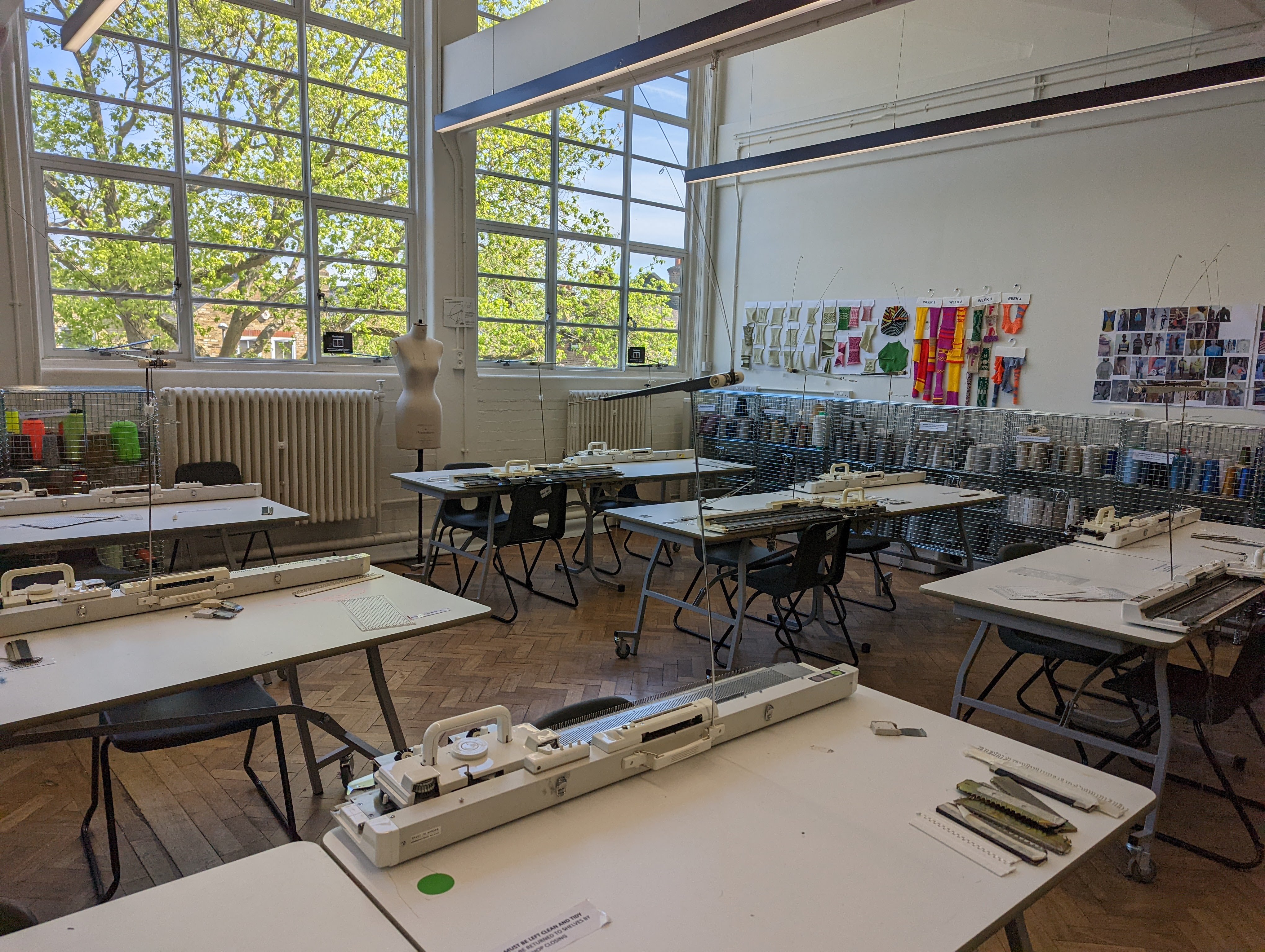 Large white room with big windows covering 1 of the walls. The room has wooden flooring and 6 tables covered in various textiles equipment. On the walls there are mood boards and coloured fabric pinned on up.