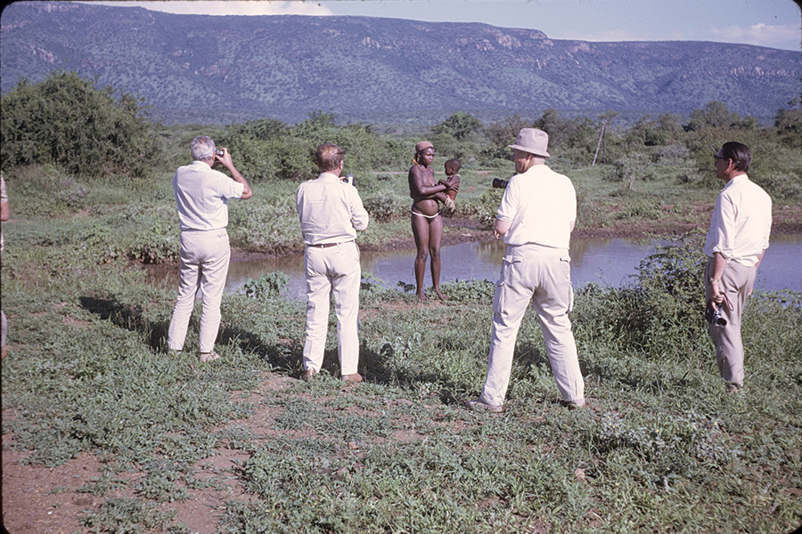 Four white men taking pictures of an African tribeswoman holding an infant by a pond in an open savanna.