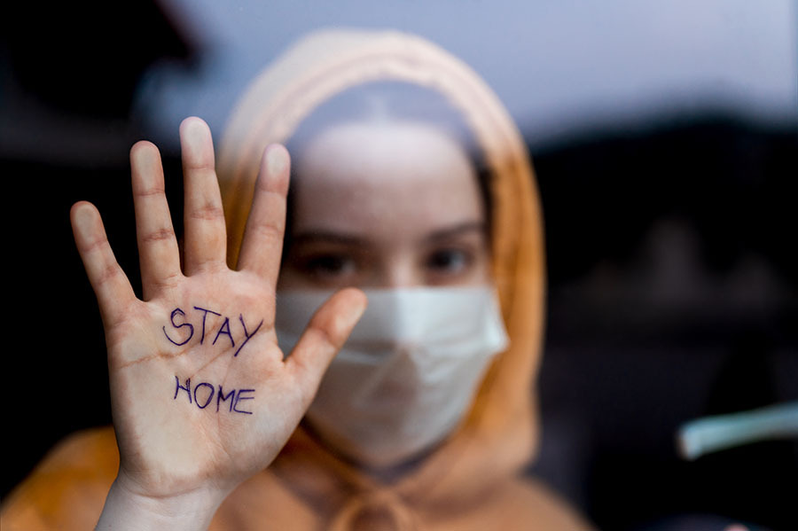 Girl with mask looks through window showing hand with text Stay home