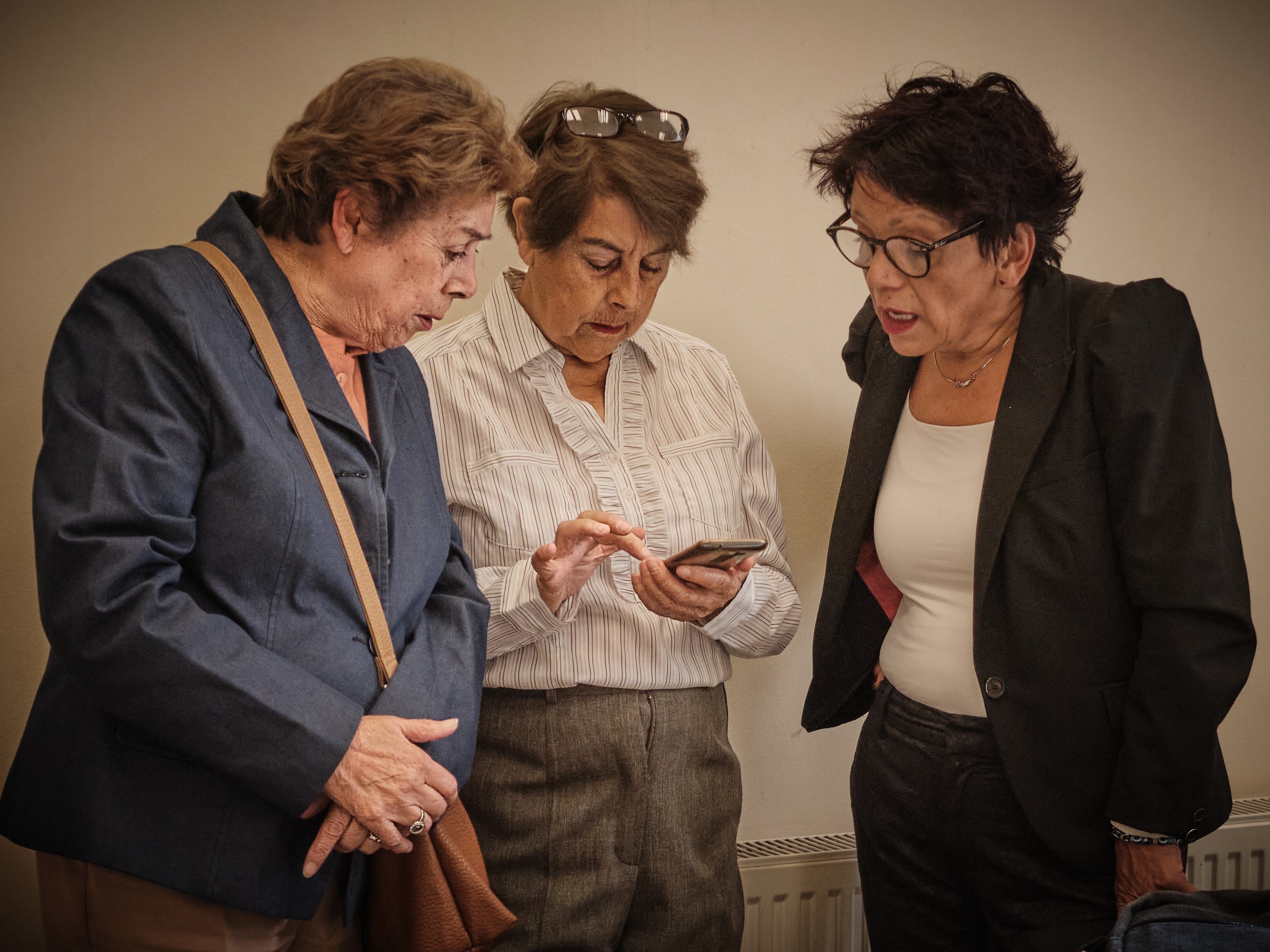 Three women dressed smartly, two in blazers and the middle one in a smart white shirt, are shown looking at a phone in a classroom in Santiago, Chile. They are all in their 50s or 60s.