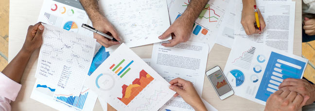 Overhead view of five sets of arms resting on a table piled with documents showing charts and graphics. 