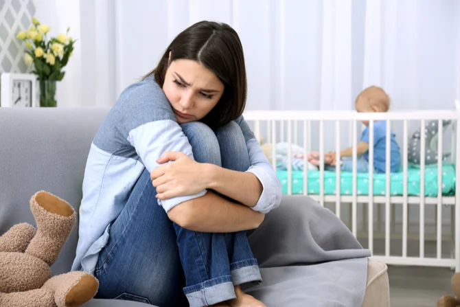 A depressed young mother sits on the floor, facing the opposite direction to her baby who sits alone in the cot.