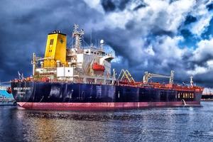 A navy blue ship in calm water near a port, under a cloudy sky