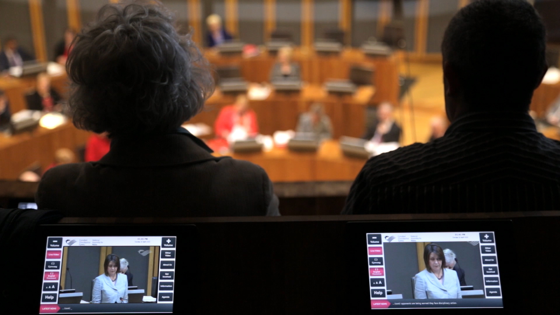 Two people sit in the chamber of the Welsh National Assembly watching politicians debate below.