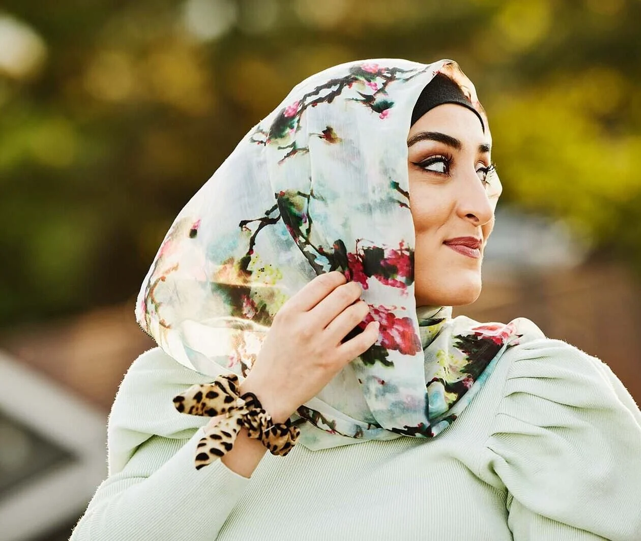 A woman is wearing a light blue patterned headscarf and a chic blue blouse with a trees in the background.