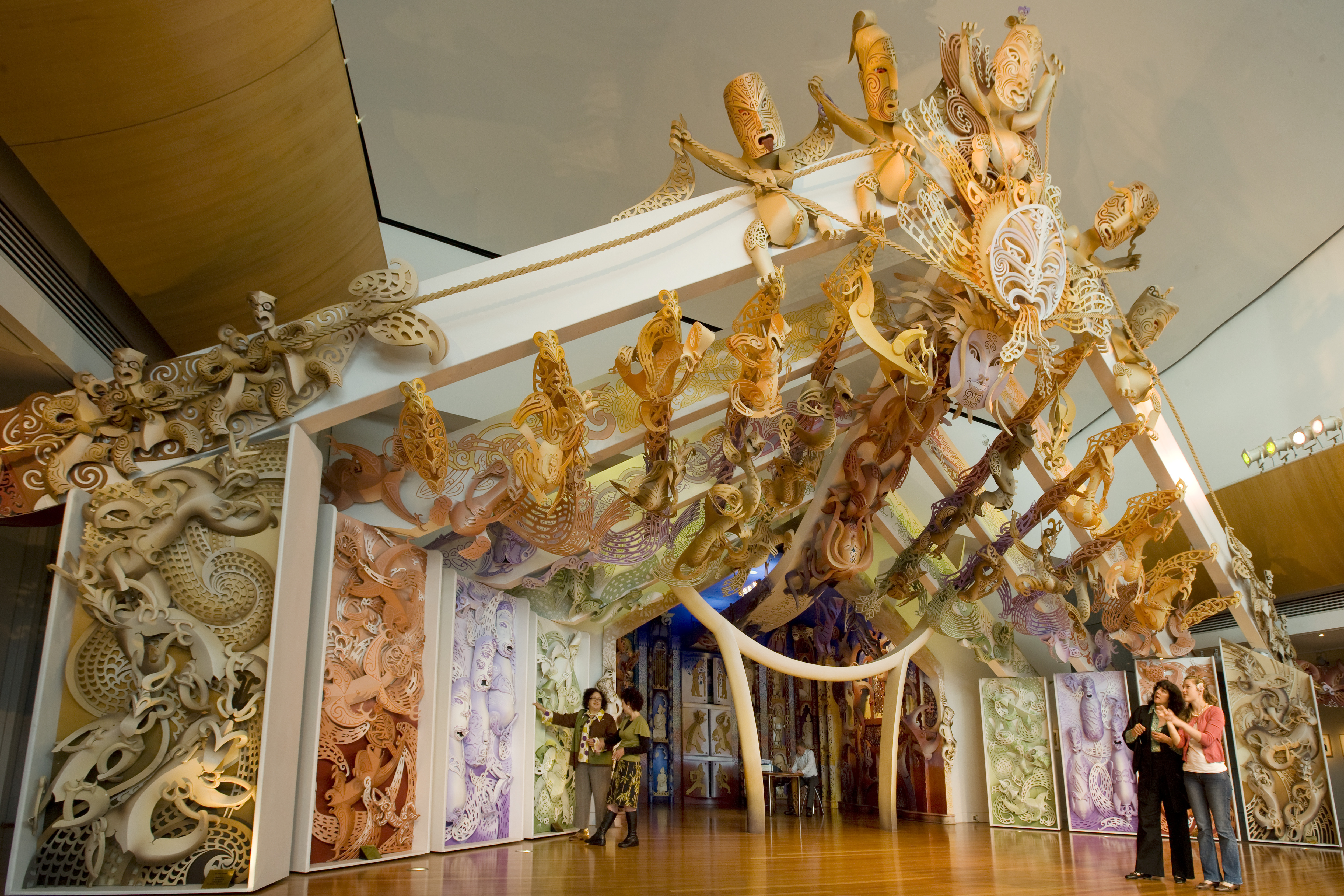 Two pairs of people admire a large carved marae (meeting house). It has many panels of intricate Māori carvings in pale colours. It is crowned with several stylized characters flanking the sun.