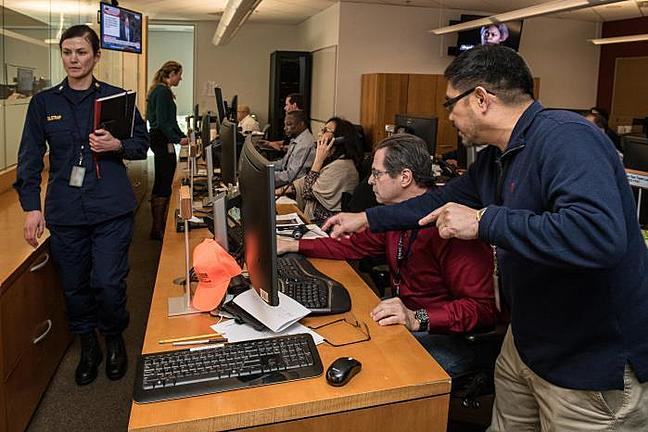 A team of 5 people sit along a desk lined with computers. One man is standing and gesturing towards the computer. A woman is also pictured walking and holding documents. These are support staff for the CDC's emergency operations center..