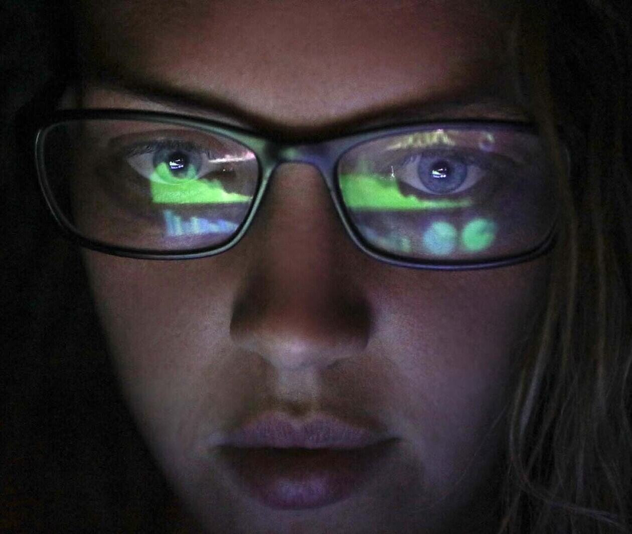 A woman with long hair stares at a computer screen reflected in her glasses, in a dark room.