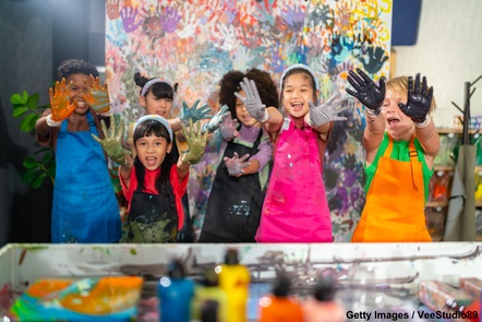 A group of six young children who are holding their paint-covered hands out towards the camera, with an out-of-focus wall covered in handprints in the background.
