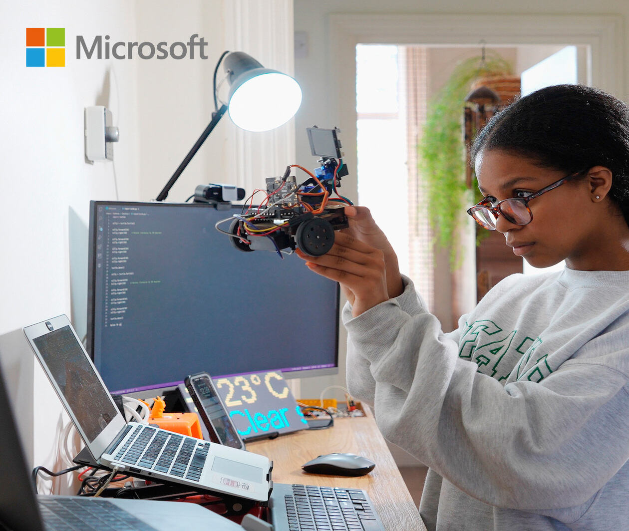 A young girl holding up a small robot she has built. Her two laptops, mobile phone, monitor and display panel are all open and active.