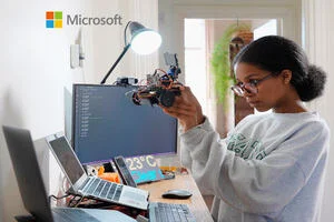 A young girl holding up a small robot she has built. Her two laptops, mobile phone, monitor and display panel are all open and active.“>
                 </div>
                 <div>
                  <div class=