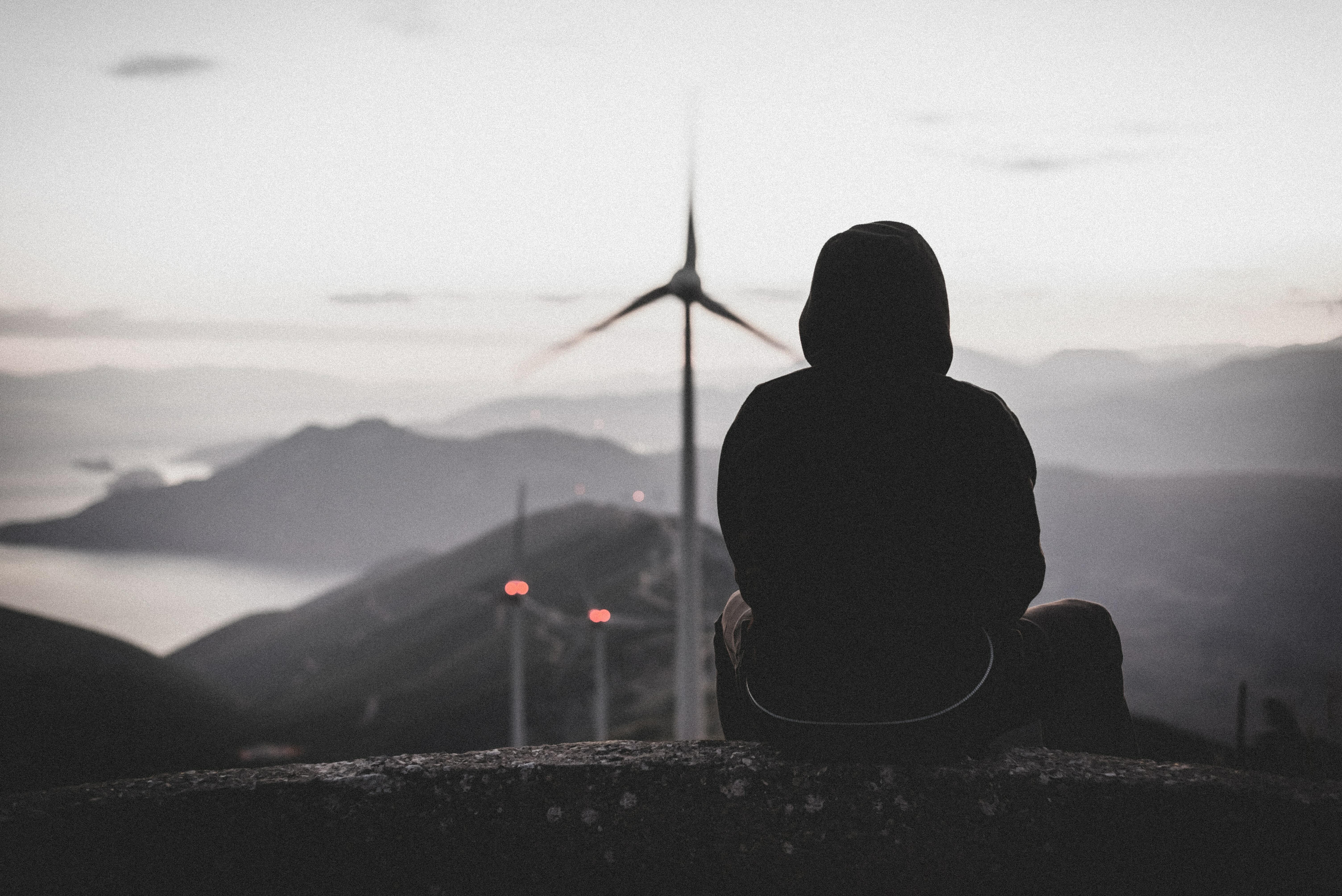 Photo of a person sitting on the edge of a building looking at a wind farm