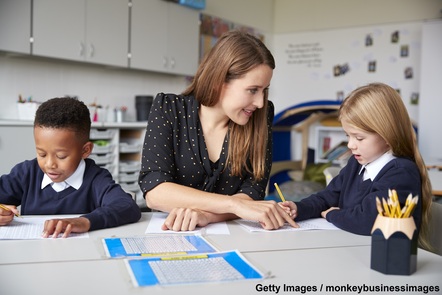 A female teacher points to a young female pupil's work. The pupil is holding a pencil of a sheet of paper. A young male pupil is holding a pencil over a piece of paper on the other side of the image.