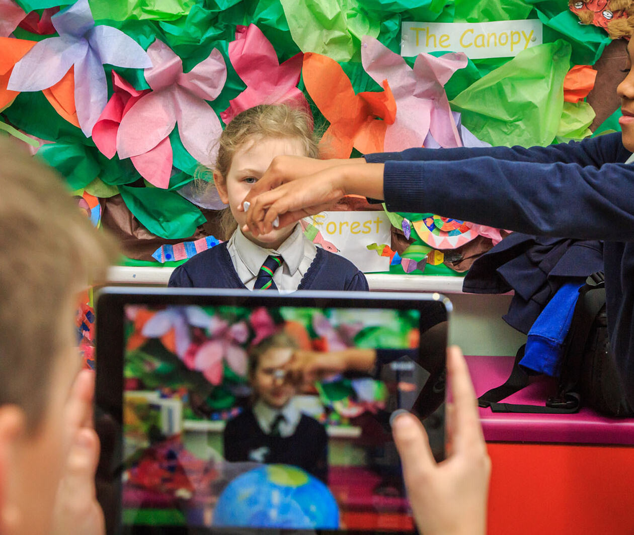 A group of three children. A girl sits as another stands to her left sprinkling paper in front of her face onto a globe. A boy is filming this on a tablet device. The background wall is decorated with colourful paper leaves and the word 'forest'.