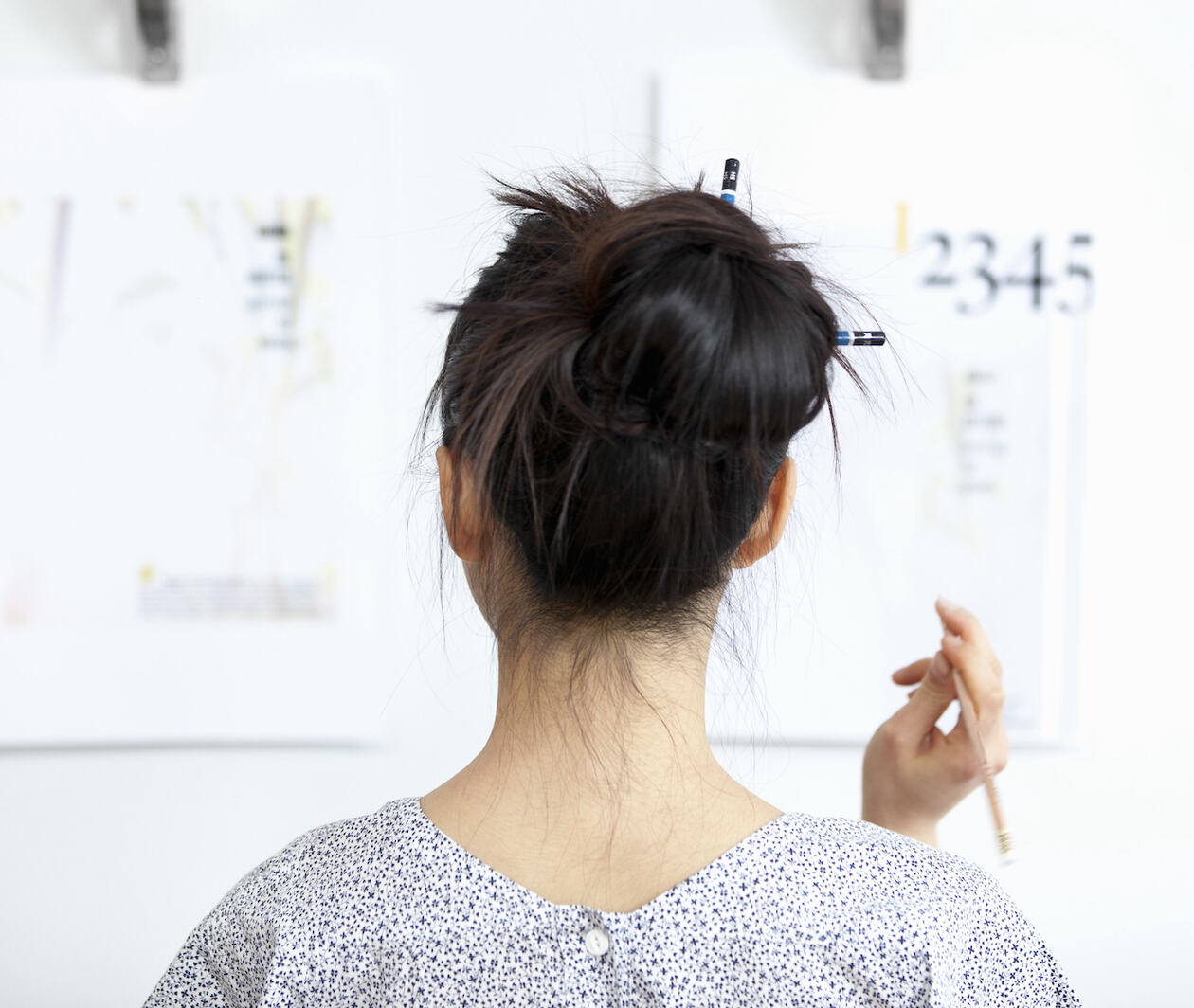 Back of a woman's head, hair in a bun with pencils in it, as she looks at a board with mathematical equations on it