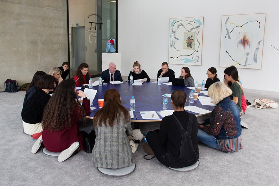 Decorative image, people sat on cushions at a large round table, reading documents on the table. Turn the Tide – Curating Contemporary Art graduate project, Show 2017