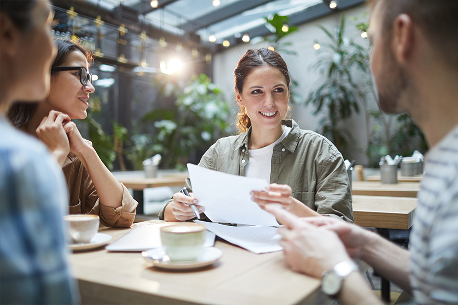 A woman holds up some papers as she and three other people smile while talking about something around a table.