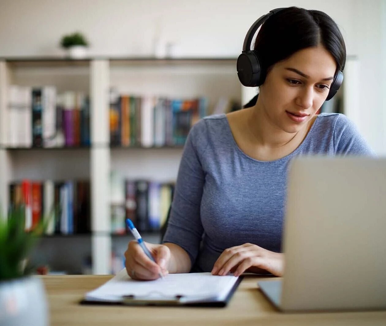 A woman sits at a desk writing in a notepad, with headphones on, looking at her laptop.