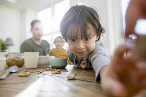 Smiling young child in foreground reaching out to the camera with a man and baby on his knee in the background. They are sitting at a breakfast table.