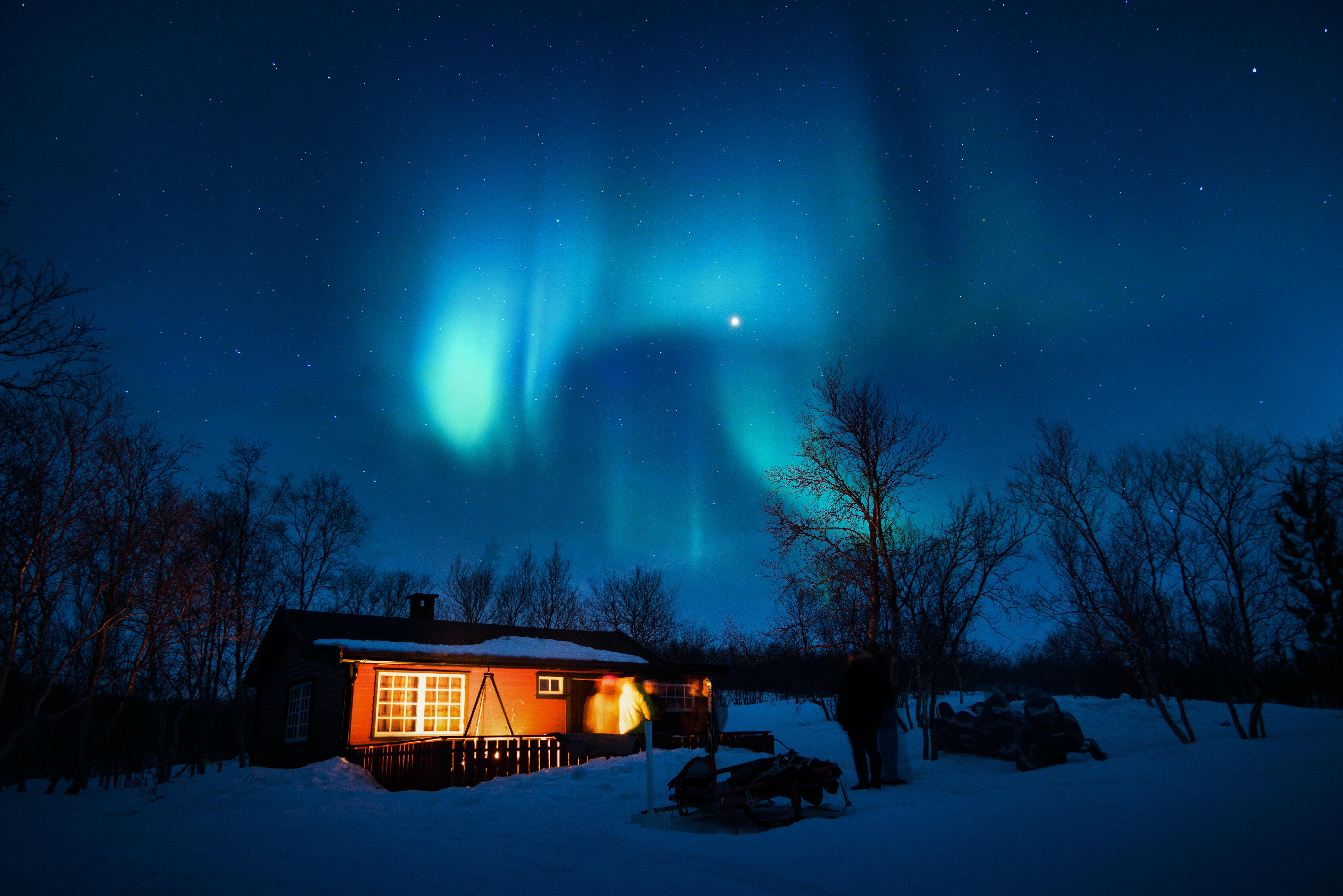 Northern lights over a night sky and a house in the snowy forest.