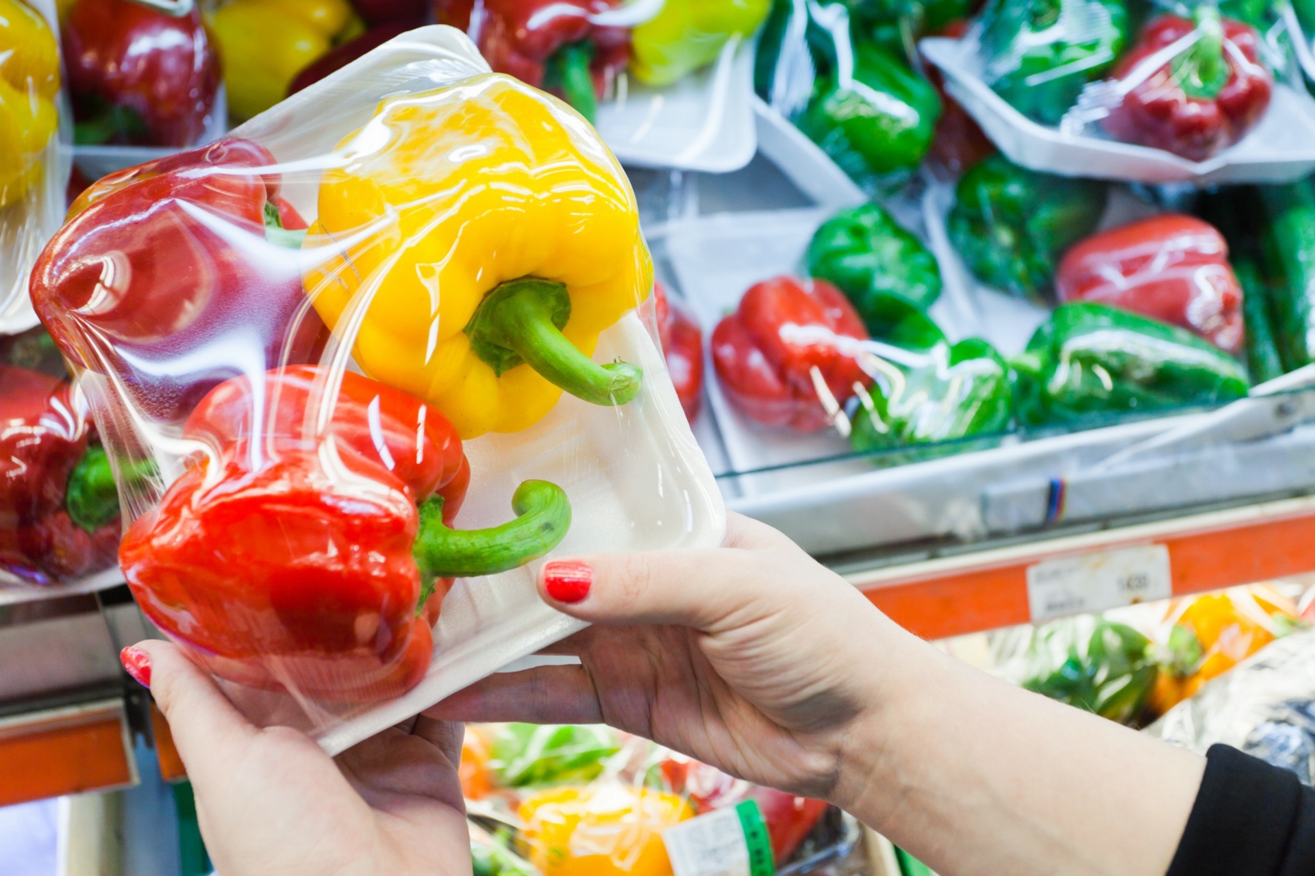 Hands holding a package of three peppers, two red, one yellow, on a white tray with plastic wrap, with similar packages in the background.