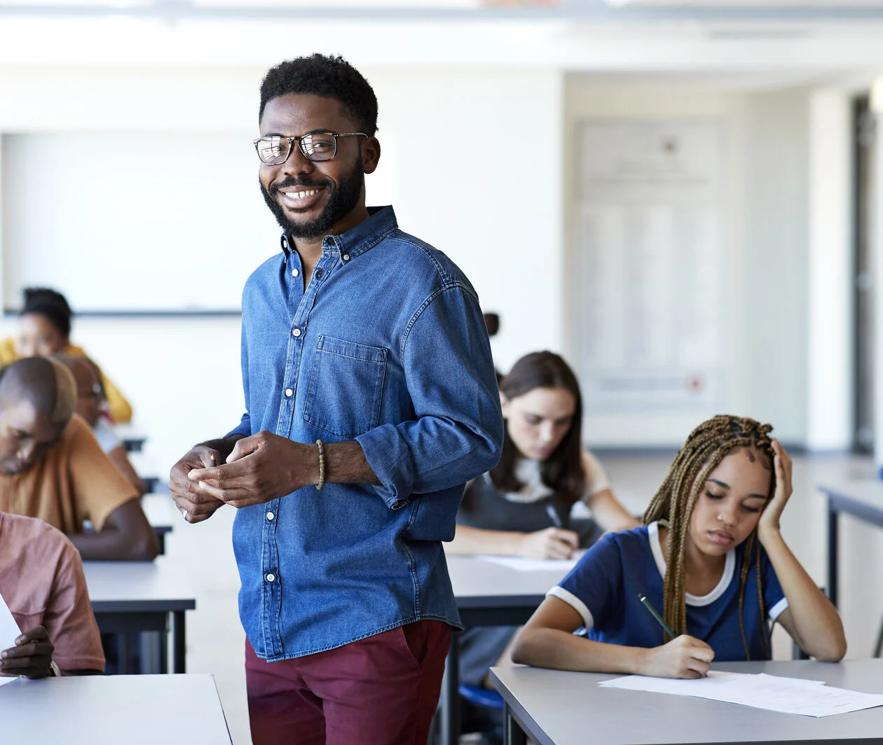 a teacher standing up in the middle of a class whilst students work on a test paper