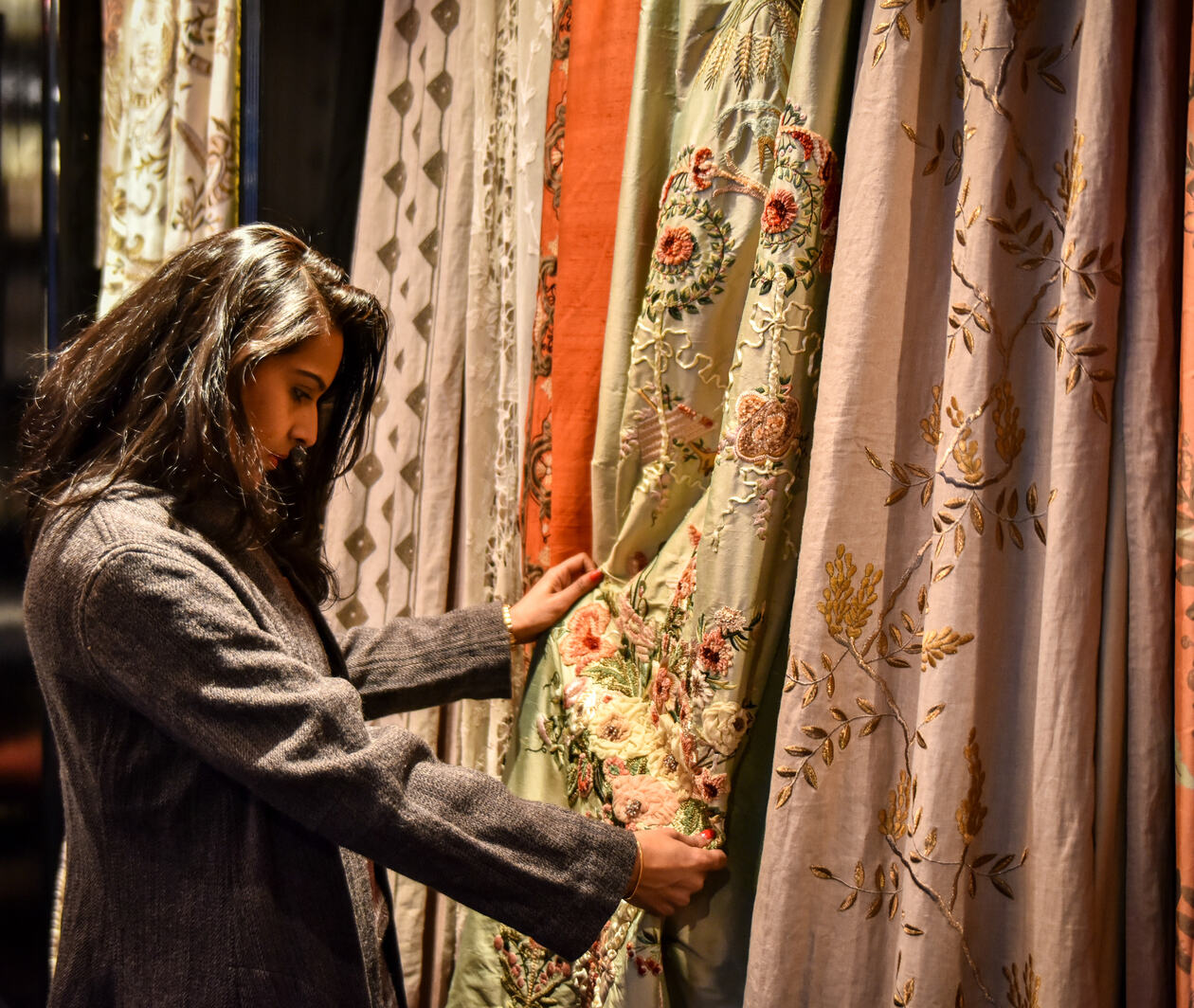 A woman in a gray jacket holds a gold floral fabric sample hanging amongst various colorful, patterned fabrics.