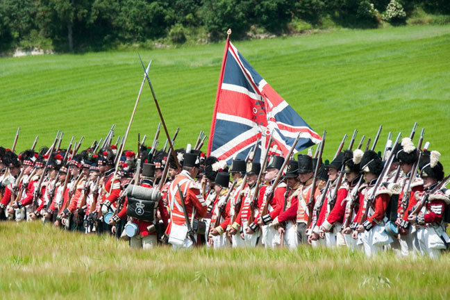 Units of the Guards in the British army practise their drill, assembled in line in a field. The Union Jack flag flies above them.