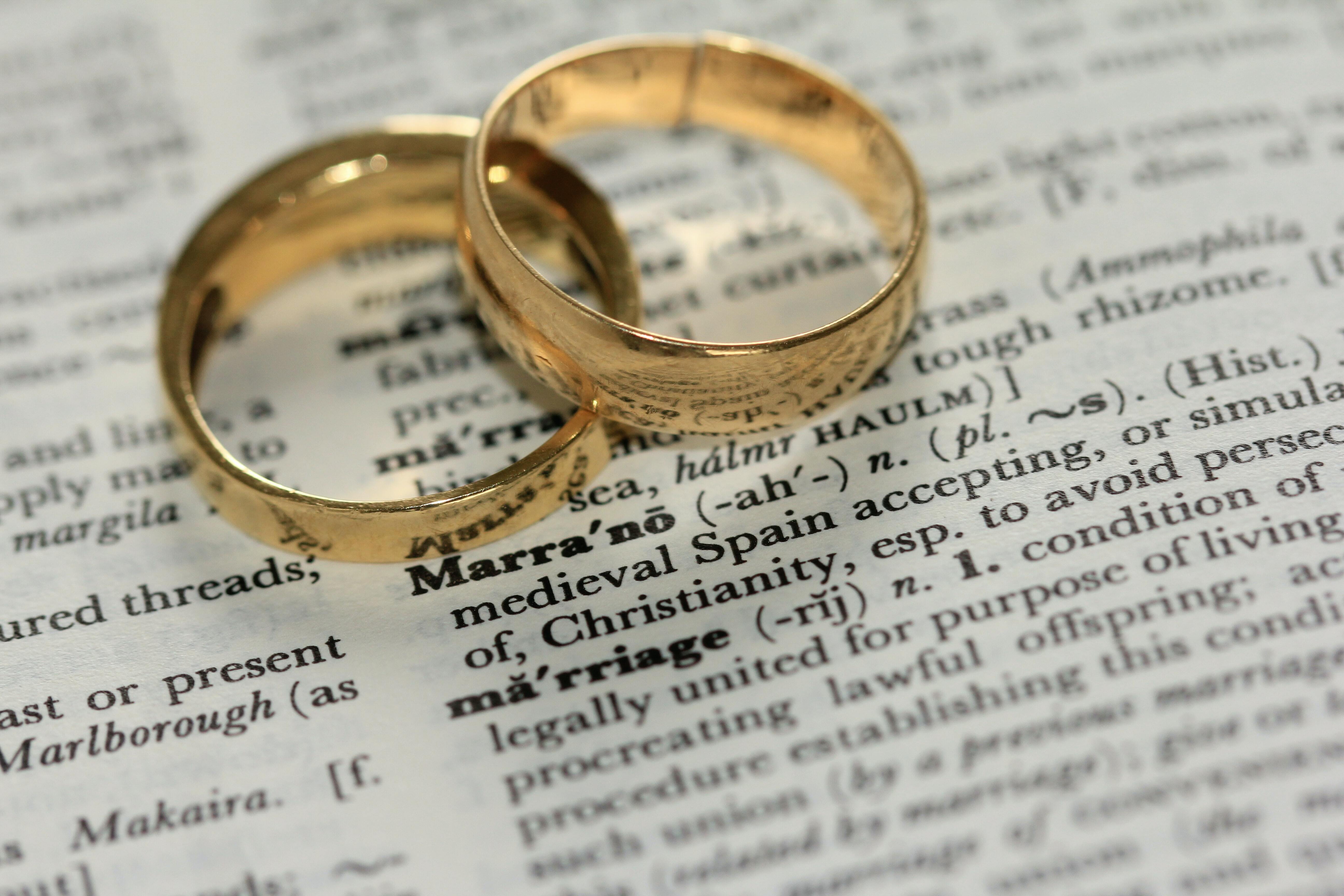 A pair of gold wedding rings placed on top of one another so they interlink. They are on top of a dictionary and the definition of 'marriage' is in focus, almost visible.