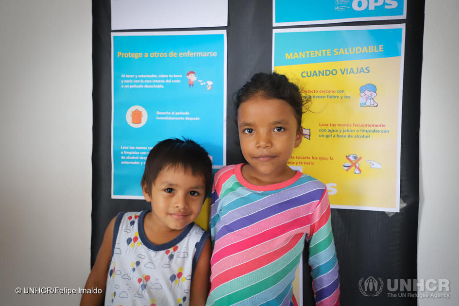 Young boy and girl smiling at the camera in front of signs explaining hygiene.