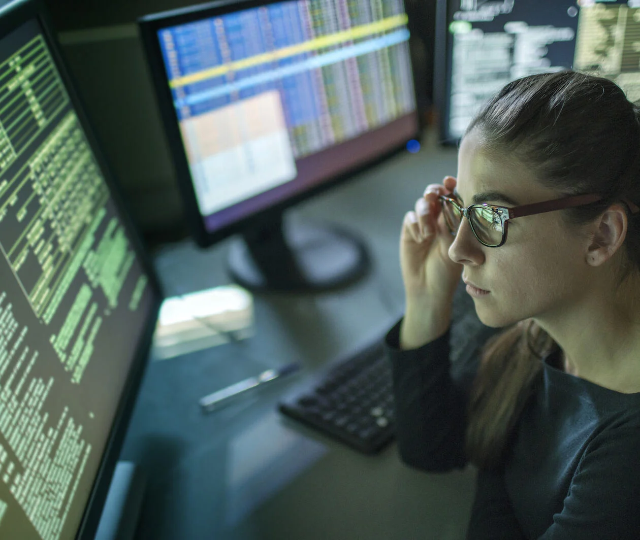 A young woman is seated at a desk surrounded by monitors displaying data