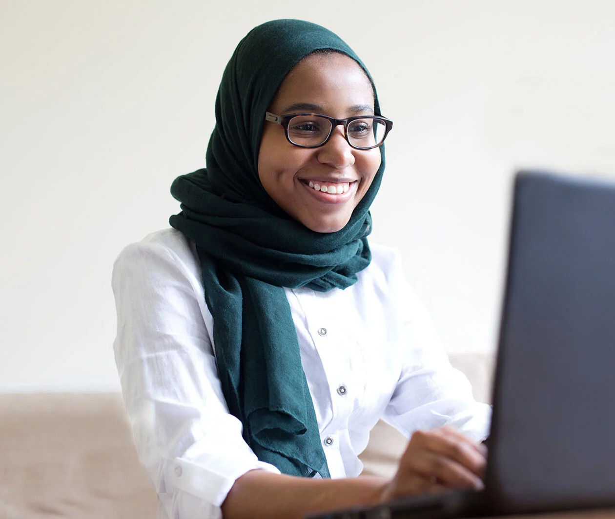 A woman in a dark blue hijab and white shirt sits on her laptop smiling. 