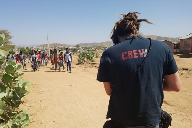 A man is visible from behind in the front of the photo. He is wearing a tshirt with CREW written on the back and has headphones on his head and carries equipment bags around his waist. He is looking towards a group of people walking along a dusty road.