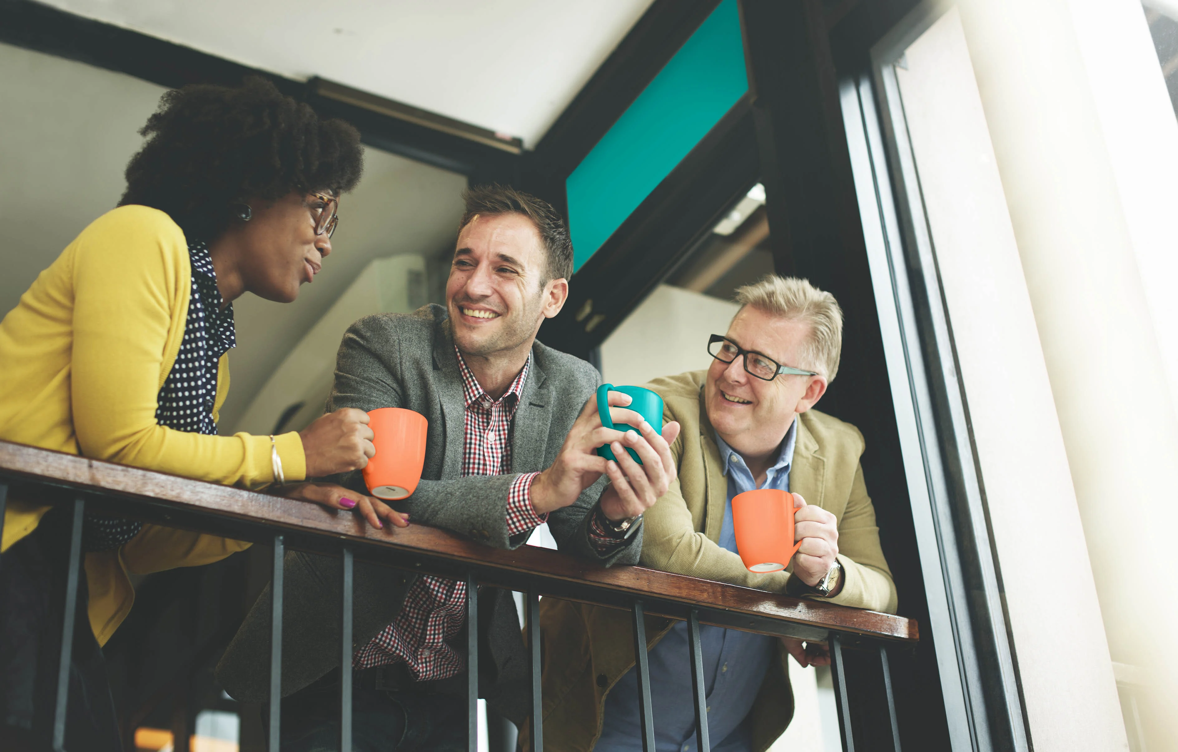 Three people talking to each other, while holding coffee mugs as they lean on a balcony railing.